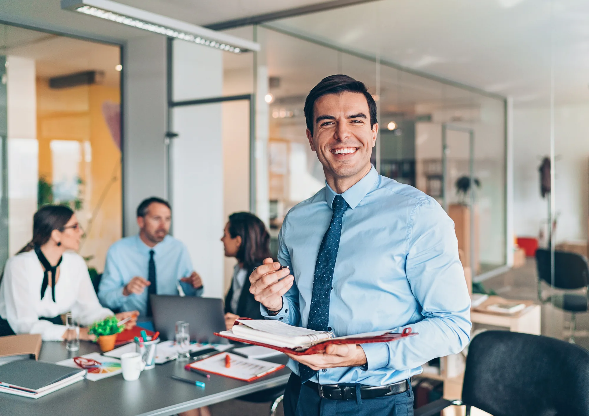 Finance-Professional im Büro, im Hintergrund Kolleginnen und Kollegen an einem Besprechungstisch